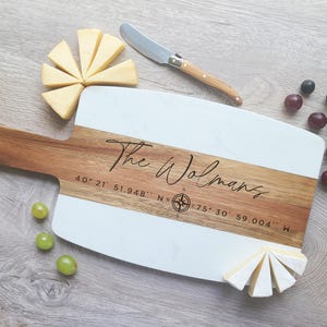 May include: A white and wood serving board with the text "The Wolmans" and coordinates "40&deg; 21' 51.948" N, 75&deg; 30' 59.004" W" engraved on it. The board is surrounded by cheese wedges and grapes.