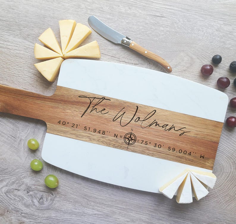 May include: A white and wood serving board with the text "The Wolmans" and coordinates "40&deg; 21' 51.948" N, 75&deg; 30' 59.004" W" engraved on it. The board is surrounded by cheese wedges and grapes.