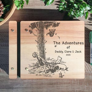 Peut inclure: Un album photo en bois avec une illustration grav&eacute;e au laser d'un arbre et de personnages. Le texte "The Adventures of Daddy, Clara & Jack 2025" est &eacute;galement grav&eacute;. Un marqueur vert est &agrave; droite.