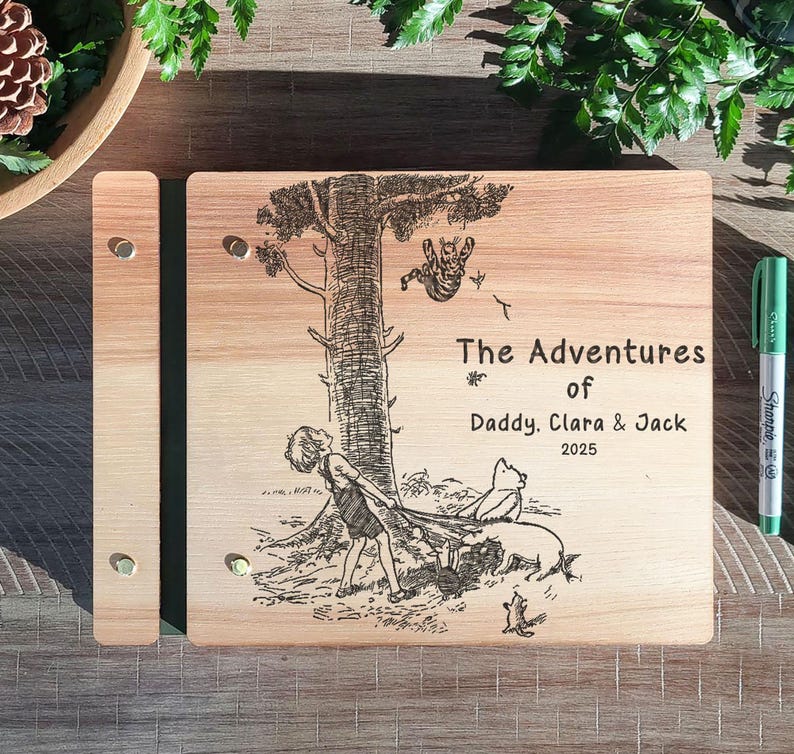 Peut inclure: Un album photo en bois avec une illustration grav&eacute;e au laser d'un arbre et de personnages. Le texte "The Adventures of Daddy, Clara & Jack 2025" est &eacute;galement grav&eacute;. Un marqueur vert est &agrave; droite.