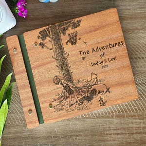 Peut inclure: Album photo en bois avec une illustration grav&eacute;e d'un arbre et de personnages. La couverture indique "The Adventures of Daddy & Levi 2025". L'album a une tranche verte et est maintenu par des rivets couleur laiton.
