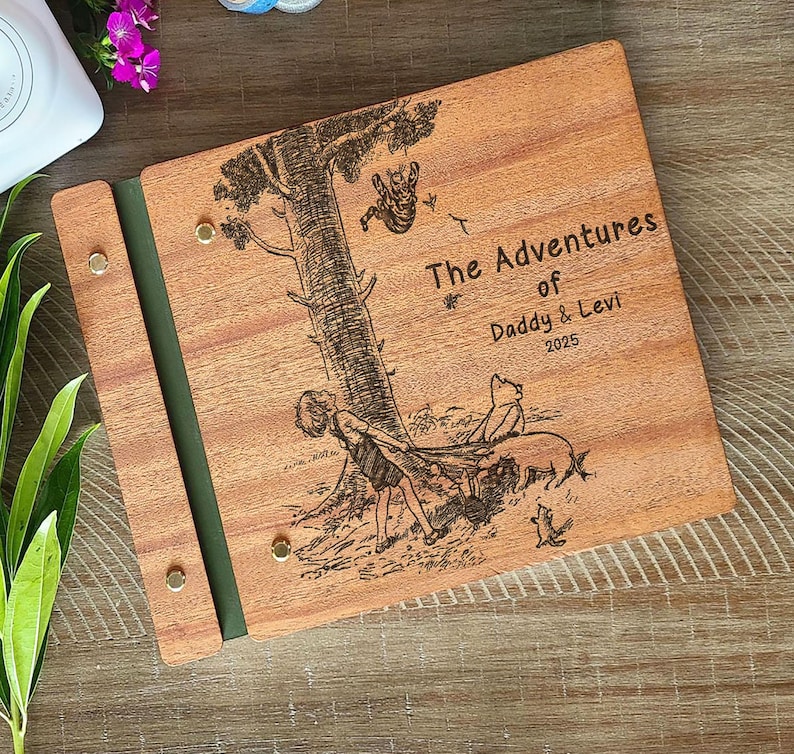 Peut inclure: Album photo en bois avec une illustration grav&eacute;e d'un arbre et de personnages. La couverture indique "The Adventures of Daddy & Levi 2025". L'album a une tranche verte et est maintenu par des rivets couleur laiton.