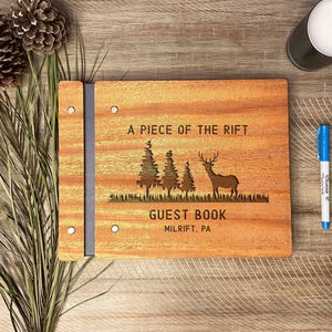 May include: Wooden guest book with the text "A PIECE OF THE RIFT" and "GUEST BOOK MILRIFT, PA". The cover features a laser-cut design of trees and a deer. Two Sharpie markers and a candle are also visible.