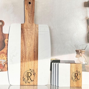 May include: A white marble and wood cutting board with a leafy design and the letter "R" engraved on the wood. The board is accompanied by a set of four white marble and wood coasters with the same design and letter.