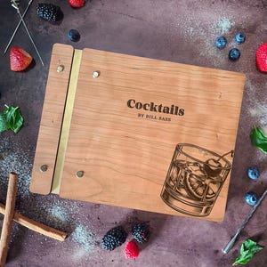 May include: A wooden cocktail recipe book with a brass spine and a vintage illustration of a cocktail glass with a cherry. The book cover is engraved with the text "Cocktails by Bill Sass".