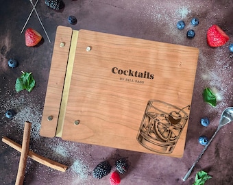 Pode incluir: Um livro de receitas de coquetéis de madeira com uma lombada de latão e uma ilustração vintage de um copo de coquetel com uma cereja. A capa do livro é gravada com o texto "Cocktails by Bill Sass".