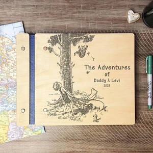 Peut inclure: Un album photo en bois avec un dos bleu. La couverture est grav&eacute;e d'une illustration en noir et blanc d'un enfant, d'un cochon et d'un ours sous un arbre. Le texte indique "The Adventures of Daddy & Levi 2025".