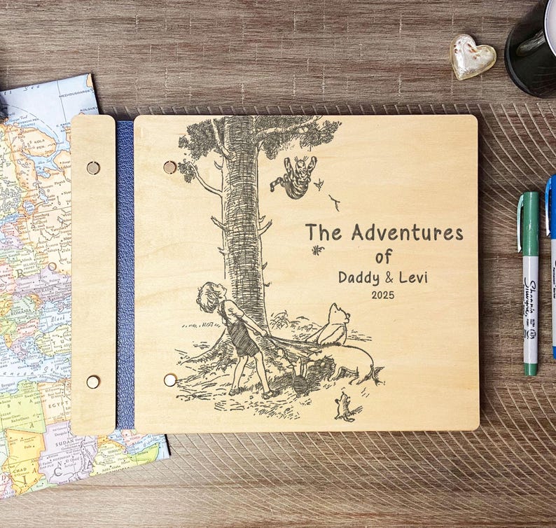 Peut inclure: Un album photo en bois avec un dos bleu. La couverture est grav&eacute;e d'une illustration en noir et blanc d'un enfant, d'un cochon et d'un ours sous un arbre. Le texte indique "The Adventures of Daddy & Levi 2025".