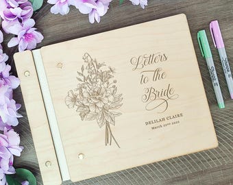 May include: A rectangular wooden guest book with a light wood grain finish, featuring an engraved floral bouquet and the text "Letters to the Bride," "DELILAH CLAIRE," and "March 22ND 2025." The book has a white spine secured with gold metal rivets.