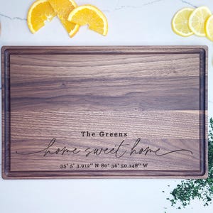 May include: A rectangular walnut wood cutting board with a personalized inscription that reads "The Greens home sweet home 35&deg; 5' 3.912" N 80&deg; 36' 50.148" W".