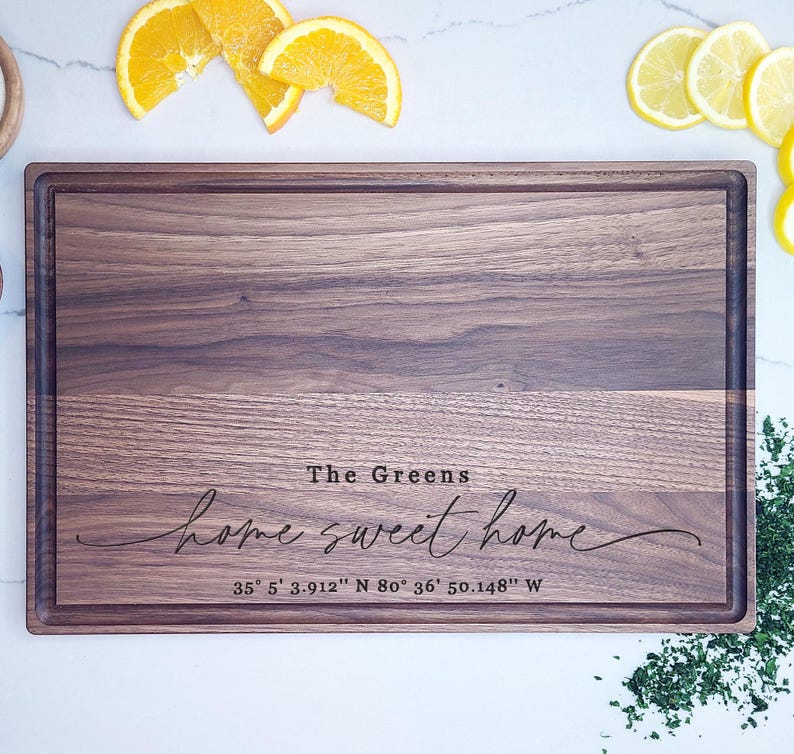 May include: A rectangular walnut wood cutting board with a personalized inscription that reads "The Greens home sweet home 35&deg; 5' 3.912" N 80&deg; 36' 50.148" W".