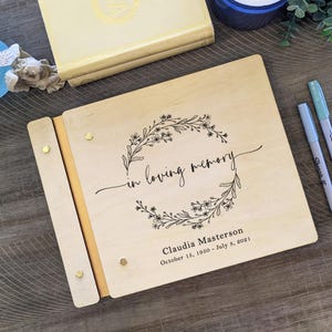 May include: A wooden photo album with a floral wreath design and the text "in loving memory". The album is personalized with the name "Claudia Masterson" and the dates "October 18, 1950 - July 8, 2021".
