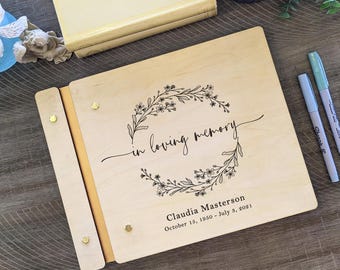 Może przedstawiać: Drewniany album ze zdjęciami z kwiatowym wiankiem i tekstem "in loving memory". Album jest spersonalizowany imieniem "Claudia Masterson" i datami "October 18, 1950 - July 8, 2021".