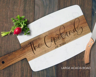 May include: A personalized acacia wood and marble serving board with the name "The Copelands" and the year "est. 2020" engraved. Includes a cheese knife and a small bunch of parsley and a radish. The board is approximately 18 inches long.