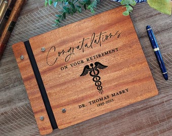 May include: A wooden guest book with a black ribbon closure. The cover is engraved with the text "Congratulations on your retirement", a medical symbol, "Dr. Thomas Mabry 1990-2025".