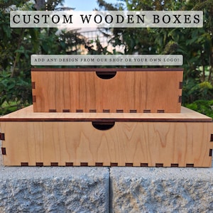 May include: Two custom wooden boxes, one atop the other, with a natural wood finish. The boxes have a dark brown trim and a curved handle cutout. The top box is smaller and a darker shade of wood. Text at the top reads "CUSTOM WOODEN BOXES".