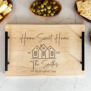 May include: A wooden serving tray with black handles, engraved with the text "Home Sweet Home", "est. 2025", "The Smiths", and "125 Mockingbird Lane". The tray features a design of three houses with a heart above them.