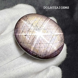 Peut inclure: Une pierre précieuse cabochon de forme ovale présentant un effet d'étoile à six branches distinct, appelé astérisme. La pierre affiche une palette de couleurs violet clair et beige translucide avec une grille d'inclusions sombres en forme d'aiguilles. Le texte indique DOLATZAIGEMS.