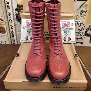 May include: A pair of red leather Dr. Martens boots with white stitching and a floral design. The boots are lace-up and have a black sole. The boots are sitting on a box with the Dr. Martens logo.