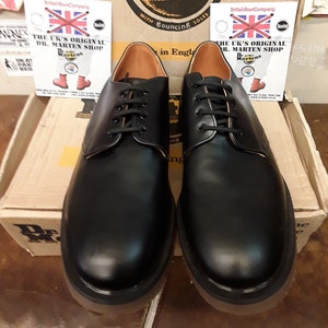 K&ouml;nnte beinhalten: Ein Paar schwarze Lederschuhe von Dr. Martens mit Schn&uuml;rsenkeln und braunem Lederfutter. Die Schuhe stehen auf einem Karton mit dem Dr. Martens Logo und dem Text "With Bouncing Soles".