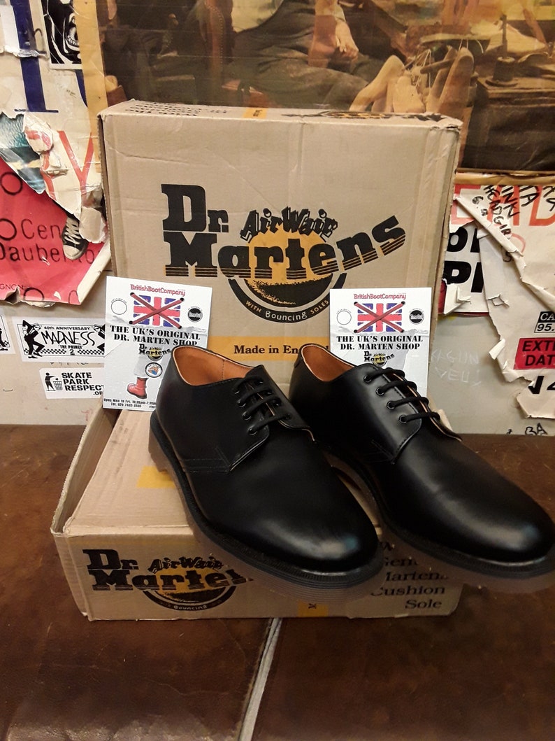 K&ouml;nnte beinhalten: Ein Paar schwarze Lederschuhe von Dr. Martens mit Schn&uuml;rsenkeln. Die Schuhe stehen auf einem braunen Karton mit dem Dr. Martens Logo. Auf dem Karton steht auch "The UK's Original Dr. Martens Shop".