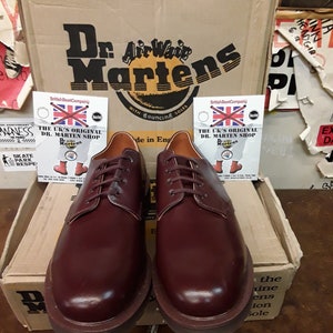Puede incluir: Un par de zapatos Oxford Dr. Martens color burdeos con cordones a juego, exhibidos frente a una caja de cart&oacute;n con el logotipo de Dr. Martens. La caja tambi&eacute;n tiene el texto "AirWair Martens" y "With Bouncing Soles".