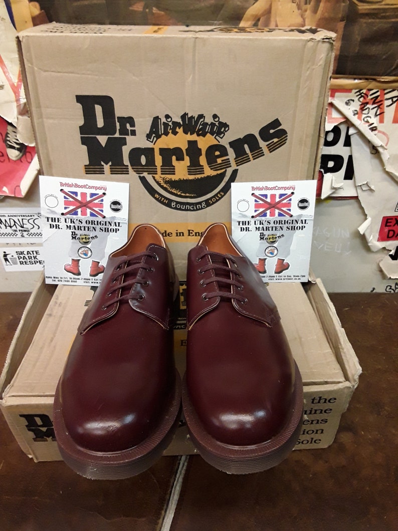 Puede incluir: Un par de zapatos Oxford Dr. Martens color burdeos con cordones a juego, exhibidos frente a una caja de cart&oacute;n con el logotipo de Dr. Martens. La caja tambi&eacute;n tiene el texto "AirWair Martens" y "With Bouncing Soles".