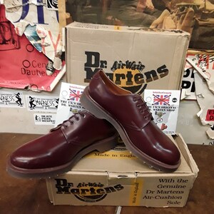 Puede incluir: Zapatos Oxford Dr. Martens burdeos con dise&ntilde;o de cordones, exhibidos sobre una caja Dr. Martens. La caja presenta el logotipo y texto de Dr. Martens. Los zapatos est&aacute;n hechos en Inglaterra.