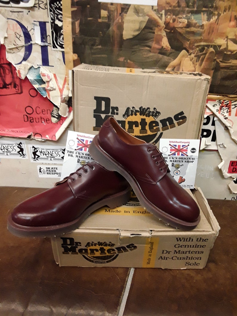 Puede incluir: Zapatos Oxford Dr. Martens burdeos con dise&ntilde;o de cordones, exhibidos sobre una caja Dr. Martens. La caja presenta el logotipo y texto de Dr. Martens. Los zapatos est&aacute;n hechos en Inglaterra.