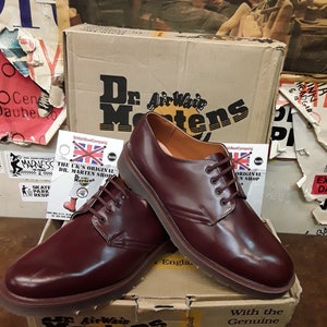 Puede incluir: Un par de zapatos Dr. Martens de cuero burdeos con cordones marrones, exhibidos sobre una caja de cart&oacute;n. La caja presenta el logotipo y el texto de Dr. Martens. Los zapatos son de estilo cl&aacute;sico, con un acabado pulido.