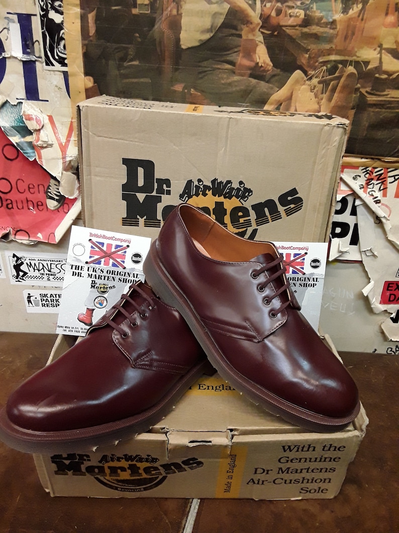 Puede incluir: Un par de zapatos Dr. Martens de cuero burdeos con cordones marrones, exhibidos sobre una caja de cart&oacute;n. La caja presenta el logotipo y el texto de Dr. Martens. Los zapatos son de estilo cl&aacute;sico, con un acabado pulido.
