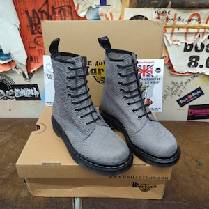 Könnte beinhalten: Ein Paar graue Lederstiefel mit schwarzen Schnürsenkeln und einem Schlangenhautmuster. Die Stiefel stehen auf einem braunen Karton mit dem Dr. Martens Logo und der Website-Adresse.