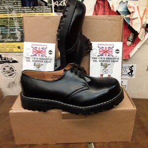 May include: A pair of black leather Dr. Martens shoes with a lace-up design and a thick, black sole. The shoes are sitting on a brown cardboard box with the text "The UK's Original Dr. Martens Shop" printed on a white label.