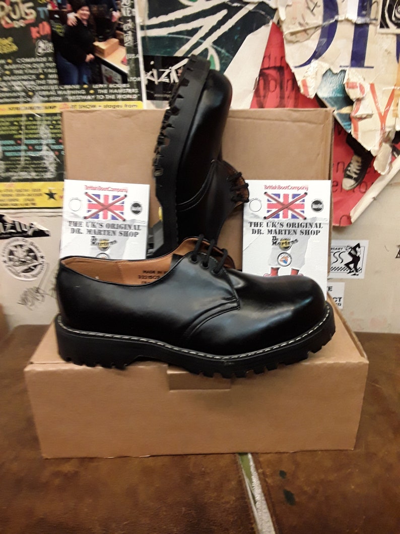 May include: A pair of black leather Dr. Martens shoes with a lace-up design and a thick, black sole. The shoes are sitting on a brown cardboard box with the text "The UK's Original Dr. Martens Shop" printed on a white label.