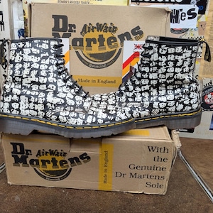 Puede incluir: Un par de botas Dr. Martens con un estampado en blanco y negro. Las botas están atadas con cordones y tienen un ribete amarillo. Las botas están encima de una caja de Dr. Martens. La caja tiene las palabras "Made in England".