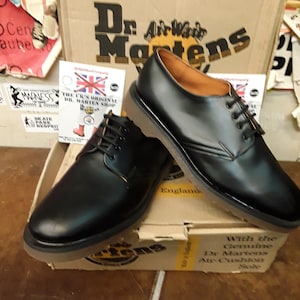 K&ouml;nnte beinhalten: Zwei Paar schwarze Lederschuhe von Dr. Martens mit dem Text "Dr. Martens" und "AirWair" auf dem Karton. Die Schuhe haben Schn&uuml;rsenkel und ein braunes Lederinnenfutter. Der Karton ist braun mit dem Text "With the Genuine Dr. Martens Air Cushion Sole" darauf gedruckt.