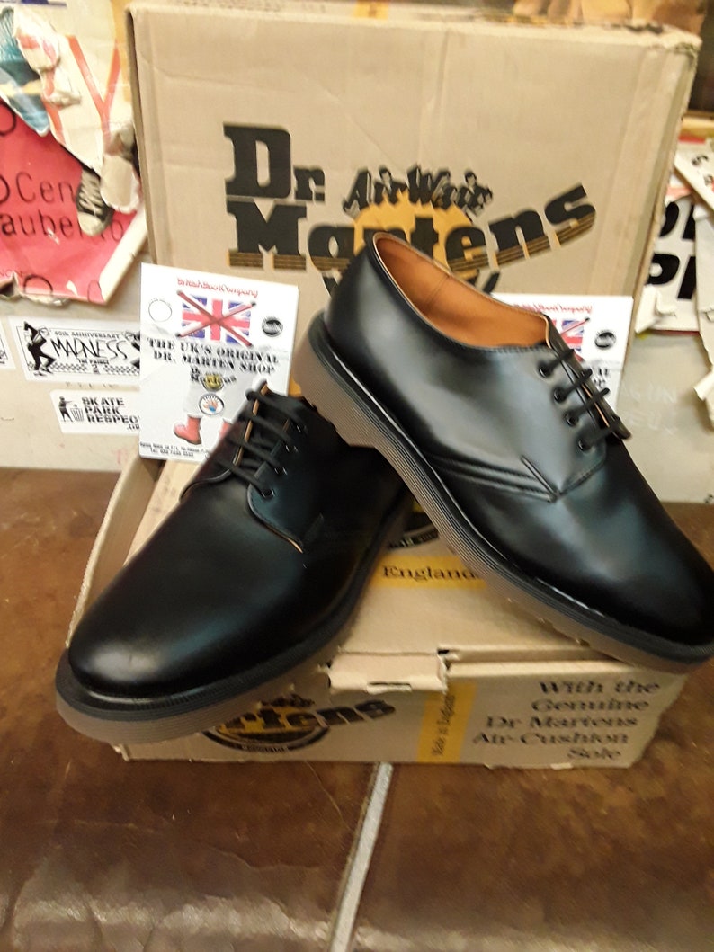 K&ouml;nnte beinhalten: Zwei Paar schwarze Lederschuhe von Dr. Martens mit dem Text "Dr. Martens" und "AirWair" auf dem Karton. Die Schuhe haben Schn&uuml;rsenkel und ein braunes Lederinnenfutter. Der Karton ist braun mit dem Text "With the Genuine Dr. Martens Air Cushion Sole" darauf gedruckt.