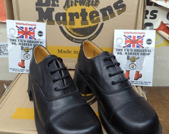 May include: A pair of black leather Dr. Martens shoes with thick soles and laces. The shoes are sitting on a box with the Dr. Martens logo and the text "Made in England" and "With Bouncing Soles".