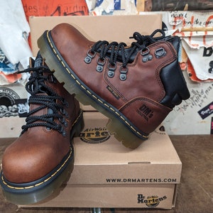 Pu&ograve; includere: Stivali in pelle marrone con lacci neri e suola in gomma nera. Gli stivali hanno il logo Dr. Martens sul lato e il testo "Anti-Static" e "DM's Industrial" sulla suola. Gli stivali sono su una scatola di cartone marrone con il logo Dr. Martens e l'indirizzo del sito web.