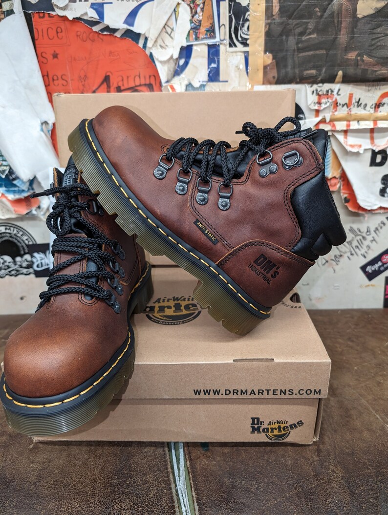 Pu&ograve; includere: Stivali in pelle marrone con lacci neri e suola in gomma nera. Gli stivali hanno il logo Dr. Martens sul lato e il testo "Anti-Static" e "DM's Industrial" sulla suola. Gli stivali sono su una scatola di cartone marrone con il logo Dr. Martens e l'indirizzo del sito web.
