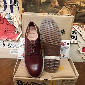 Puede incluir: Un par de zapatos borgo&ntilde;a Dr. Martens, uno mostrando la suela, sobre una caja de cart&oacute;n. La caja presenta el logotipo y texto de Dr. Martens. Los zapatos est&aacute;n atados con cordones y muestran las costuras distintivas de la marca.