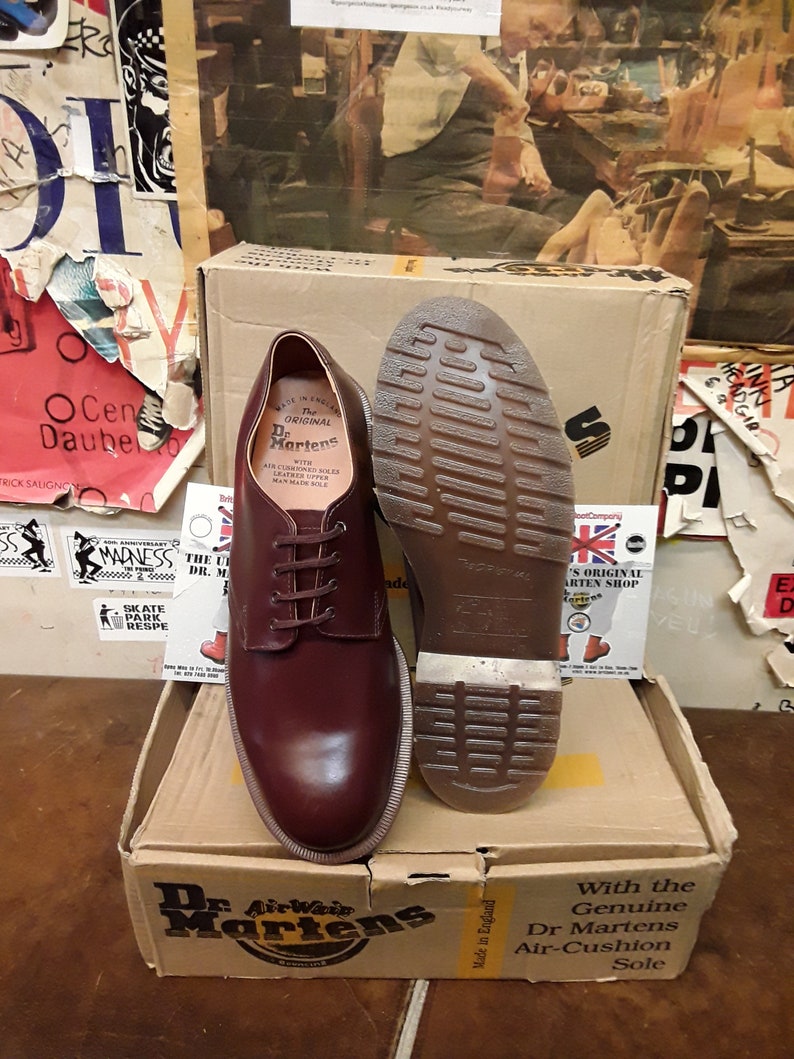 Puede incluir: Un par de zapatos borgo&ntilde;a Dr. Martens, uno mostrando la suela, sobre una caja de cart&oacute;n. La caja presenta el logotipo y texto de Dr. Martens. Los zapatos est&aacute;n atados con cordones y muestran las costuras distintivas de la marca.