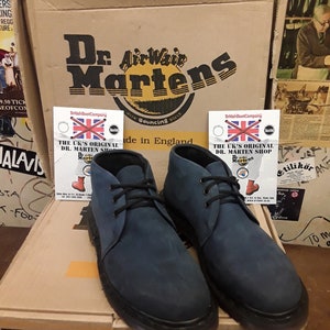 May include: A pair of dark blue suede Dr. Martens shoes with a brown box in the background. The box has the Dr. Martens logo and the text "With Genuine Dr. Martens Air-Cushion Sole" on it.