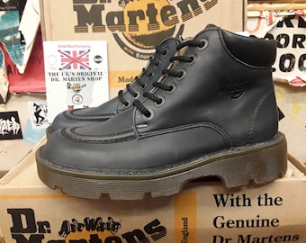 May include: Black leather Dr. Martens ankle boots with a lace-up closure and a thick, textured sole. The boots are sitting on a box with the text "Dr. Martens" and "With the Genuine Dr. Martens" printed on it.