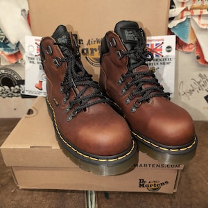 Pu&ograve; includere: Un paio di stivali Dr. Martens in pelle marrone con lacci neri e cuciture gialle. Gli stivali hanno una punta in acciaio e sono appoggiati su una scatola di cartone marrone con il logo Dr. Martens.