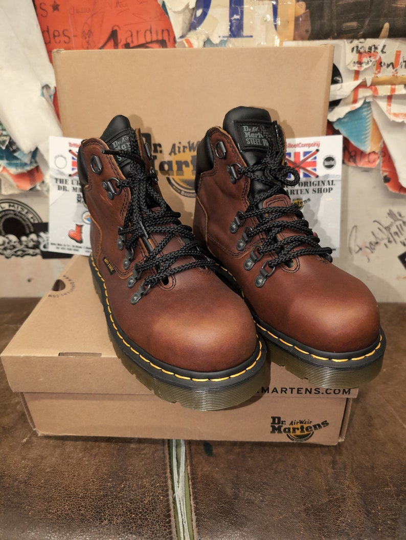 Pu&ograve; includere: Un paio di stivali Dr. Martens in pelle marrone con lacci neri e cuciture gialle. Gli stivali hanno una punta in acciaio e sono appoggiati su una scatola di cartone marrone con il logo Dr. Martens.