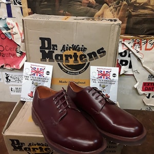 Puede incluir: Un par de zapatos Oxford Dr. Martens burdeos con cordones a juego, expuestos sobre una caja de cart&oacute;n con el logotipo de la marca. La caja y los zapatos est&aacute;n frente a una pared cubierta de carteles. Fabricado en Inglaterra.