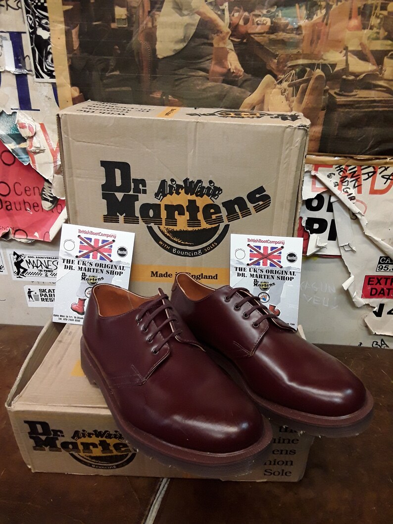 Puede incluir: Un par de zapatos Oxford Dr. Martens burdeos con cordones a juego, expuestos sobre una caja de cart&oacute;n con el logotipo de la marca. La caja y los zapatos est&aacute;n frente a una pared cubierta de carteles. Fabricado en Inglaterra.
