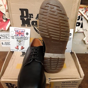 K&ouml;nnte beinhalten: Ein Paar schwarze Lederschuhe von Dr. Martens mit Schn&uuml;rung und einer dicken, strukturierten Sohle. Die Schuhe stehen auf einem braunen Karton mit dem Dr. Martens Logo. Auf dem Karton steht auch "With the Genuine Dr Martens Air-Cushion Sole".