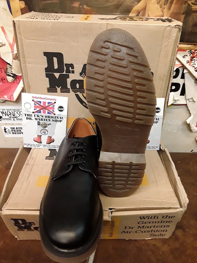 K&ouml;nnte beinhalten: Ein Paar schwarze Lederschuhe von Dr. Martens mit Schn&uuml;rung und einer dicken, strukturierten Sohle. Die Schuhe stehen auf einem braunen Karton mit dem Dr. Martens Logo. Auf dem Karton steht auch "With the Genuine Dr Martens Air-Cushion Sole".